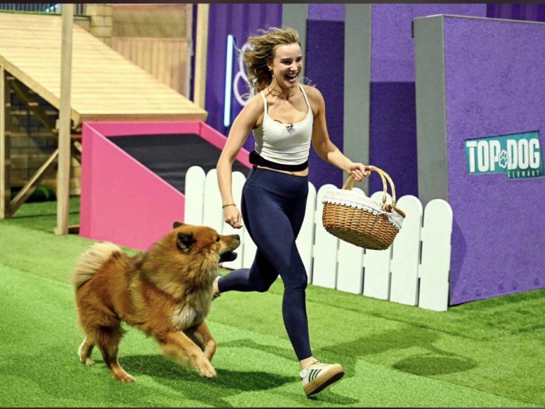 Eine Frau läuft mit einem Hund in einem Trainingsparcour, trägt einen Korb und lächelt.
