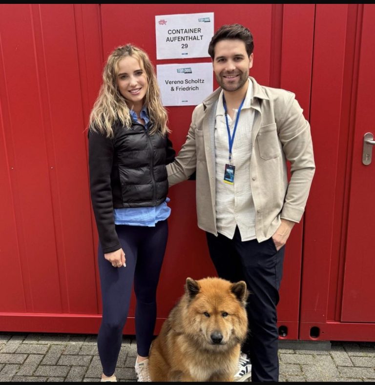 Paar mit einem Hund (Eurasier) vor einem roten Gebäude, beide lächeln in die Kamera.