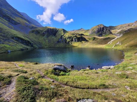 Bergsee umgeben von grünen Wiesen und steilen Hügeln unter blauem Himmel.